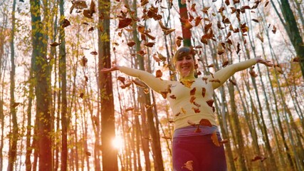 Wall Mural - SLOW MOTION, CLOSE UP, LENS FLARE, PORTRAIT: Happy young woman exploring the autumn colored woods throws the dry fallen leaves high into the air at golden sunrise. Carefree girl plays with dry leaves.