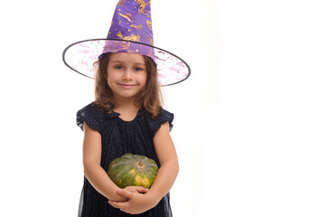 Wall Mural - Halloween Witch concept - Portrait of little Caucasian girl dressed in stylish carnival attire and wizard hat, witch child smiles posing with pumpkin against a white background with copy space