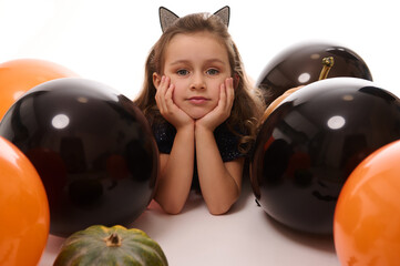 Wall Mural - Beautiful child girl with hoop with cat ears looks at camera lying down next to pumpkin, colored black and orange balloons on a white background with copy space. Halloween concept