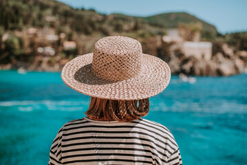 Back view of woman in striped cruise outfit, straw hat. Lady standing on stone beach. Teal sea background. South Europe Riviera, travel destination, luxury vacation concept.