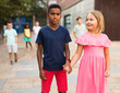 Cheerful cute preteen girl walking with her african american friend along city street on summer day ..