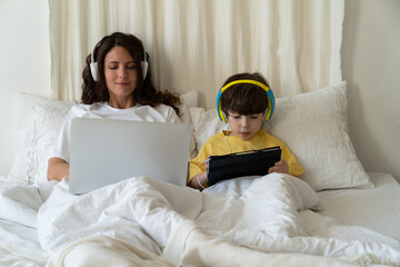 Wall Mural - Work and education on lockdown: mother type on laptop computer lying in bed together with preschool son kid watch educational video or cartoon on digital tablet. Family routine at home on quarantine