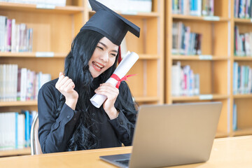 Wall Mural - Young Asian university woman graduate in graduation gown and mortarboard with degree certificate celebrates with family through video call on laptop in virtual convocation during COVID-19 pandemic