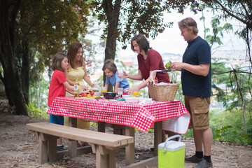 Smiling family setting table for picnic. Mother, father and children getting ready for meal at open air, putting food and appliances at table. Family time, outdoor activity, picnic concept