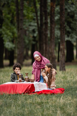 Wall Mural - Smiling kids holing tea in glasses near muslim mother in hijab in park