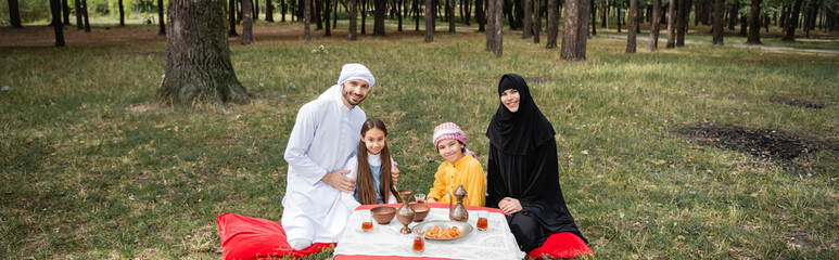 Wall Mural - Smiling muslim family looking at camera near food in park, banner