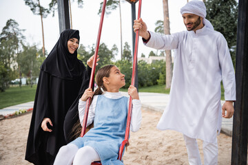 Canvas Print - Smiling muslim parents standing near daughter on swing outdoors