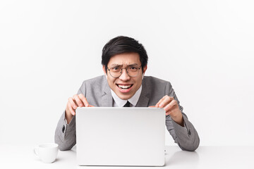 Portrait of displeased, skeptical and judgemental asian businessman in wireless earbuds, sitting desk, pointing at laptop screen with disappointed upset expression, white background