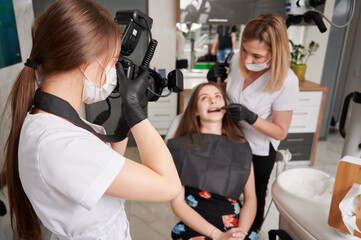 Wall Mural - Dentist examining woman teeth while assistant in medical mask taking photo with professional camera. Young female doctor in sterile gloves photographing patient and dentist in dental office.