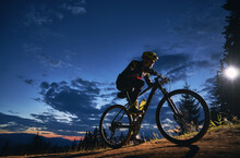 Young Man Cycling Bicycle Under Beautiful Night Sky. Male Bicyclist In Safety Helmet Riding On Hillside Road Under Blue Cloudy Sky At Night. Concept Of Sport, Biking And Active Leisure.
