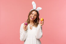 Holidays, Spring And Party Concept. Cheerful Good-looking Blond Woman Celebrating Easter Day In Rabbit Ears, Holding Two Painted Eggs And Wink Camera, Smiling Happily, Pink Background