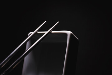 Stainless steel chopsticks on white square bowl on black background.