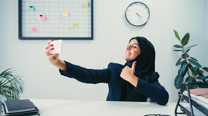 Wall Mural - happy muslim businesswoman showing thumb up while taking selfie on smartphone in office