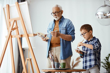 Senior Artist And His Small Grandson Use Artist's Pallete And Paint On Canvas Together.