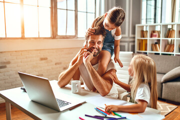 The businessman dad tries to work at the laptop when his little daughters play, fool around and interfere with him. Freelance, work from home.