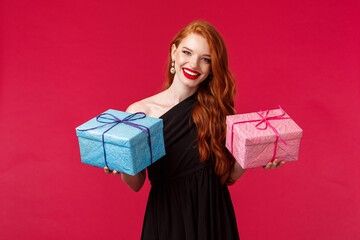 Wall Mural - Celebration, holidays and women concept. Portrait of elegant gorgeous young redhead woman in black party dress, holding two gifts in blue and pink boxes, smiling camera cheerful, red background