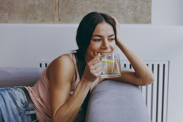 Wall Mural - Vivid young latin woman 20s in casual clothes hands folded under head sit lay on sofa hold drink water with lemon look aside spend time in living room home in own house be lost in reverie good day