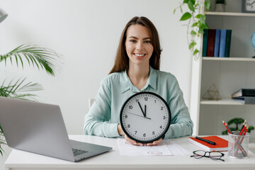 Young smiling happy fun successful employee business woman in blue shirt hold in hands clock sit work at workplace white desk with laptop pc computer at light office indoor Achievement career concept