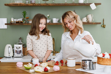 Happy chef cook baker mom woman in white shirt work with obedient child baby girl helper pouring milk at kitchen table home. Cooking food process concept Mommy little kid daughter prepare fruit cake