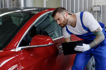 Young minded male professional car mechanic man wears denim blue overalls white t-shirt gloves estimate hold clipboard with papers document work in light modern vehicle repair shop workshop indoors