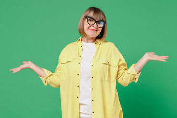 Wall Mural - Elderly smiling happy woman 50s in glasses yellow shirt shrugging shoulders looking puzzled, spread hands do oops gesture isolated on plain green background studio portrait People lifestyle concept