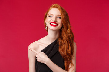 Wall Mural - Close-up portrait of delighted pretty romantic redhead woman in red lipstick, black dress, contemplate something beautiful, pointing and looking left with happy silly smile, red background