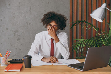 Young tired exhausted sad sick african american employee business man in classic shirt tie close cover eyes with hand sit work at white office desk with pc laptop inside Achievement career concept.