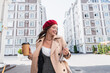 joyful woman in red beret and beige trench coat holding laptop and paper cup of street