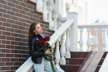 Pretty Woman In Leather Jacket And Jeans Holding Bouquet Of Red Roses On Urban Street In Europe