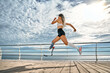 © HBS - Full length portrait of a confident young disabled sportswoman running fast on the bridge near the sea.