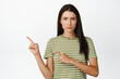 © Cookie Studio - Concerned brunette woman pointing left, frowning and looking skeptical, standing over white background