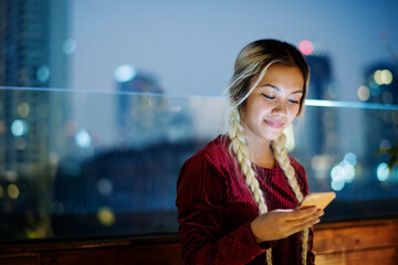 Wall Mural - Smiling young woman using a smartphone in the evening cityscape