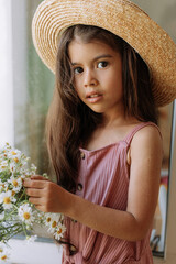 cute little swarthy brunette girl in a straw hat and a pink dress with a bouquet of daisies. purity 