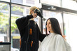 © Siam Stock - Caucasian female client in haircut cape sitting on hairdressing chair looking in the mirror and smiling while getting a hair blowing with a hairdryer by a blond Asian female hairdresser in a face mask
