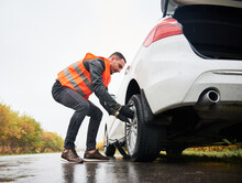 Roadside Assistance Worker Removing Flat Tire While Repairing Car On The Street. Male Auto Mechanic In Work Vest Changing Flat Tire On The Road. Concept Of Emergency Road Service.