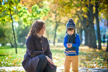 Wall Mural - Mother and baby play in autumn park. Parent and child walk in the forest on a sunny fall day. Children playing outdoors with yellow maple leaf. Toddler boy play with golden leaves. Mom hugging kid