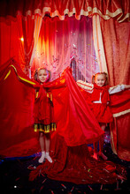 Small Girls During A Stylized Theatrical Circus Photo Shoot In A Beautiful Red Location. Young Models Posing On Stage With Curtain. Twin Sisters Or Female Friends Together