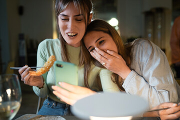 Wall Mural - Best friends watching funny content on the smartphone screen and having fun while they're eating at Chinese restaurant. Concept of girls having fun using tech.