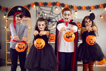 Kids Are Ready For Tricks Or Treats On Halloween Night. Four Children Standing In Room In Carnival Costumes And Spooky Make-up Stretch Their Buckets Pumpkins To Camera. Halloween Celebration Concept.