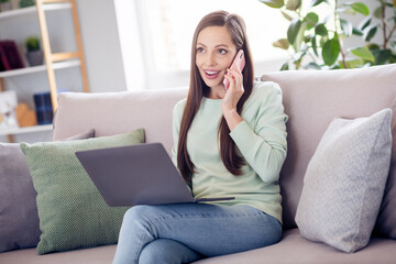 Sticker - Photo of cheerful millennial brunette lady sit on sofa with laptop talk telephone wear blue shirt at home alone