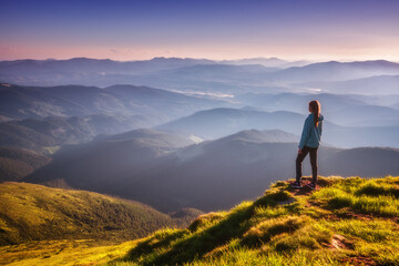 Wall Mural - Girl on mountain peak with green grass looking in beautiful mountain valley in fog at sunset in autumn. Landscape with sporty young woman, foggy hills, forest, sky in fall. Travel and tourism. Hiking