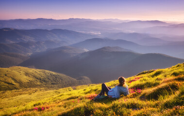 Wall Mural - Young woman on the mountain peak with green grass and pink flowers looking at beautiful mountains in fog at sunset in summer. Colorful landscape with lying girl, forest, hills, sky. Travel and tourism