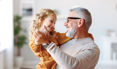 Adorable child girl and positive grandpa holding hands while dancing together in living room