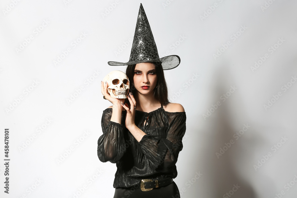 Young witch with human skull on light background