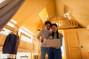Wall Mural - Man with cup hugging asian girlfriend pointing at laptop in glamping house