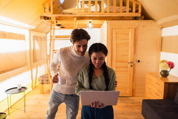 Wall Mural - Interracial couple with cup using laptop in glamping house during weekend