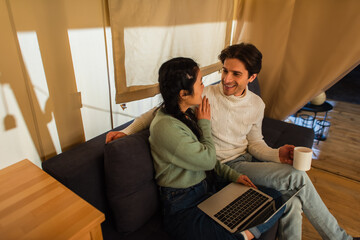 Wall Mural - Asian woman with laptop talking to boyfriend with cup in glamping house