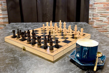 Chess Board With Figures And Blue Cup With Cappuccino Coffee On Marble Table Against Brown Curtain And Brick Wall Background