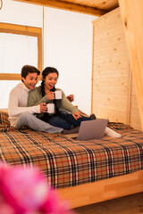Wall Mural - Excited asian woman pointing at laptop near boyfriend with cup on bed in glamping house