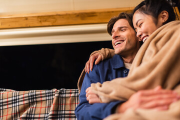 Wall Mural - Low angle view of laughing interacial couple in blanket sitting on bench outdoors